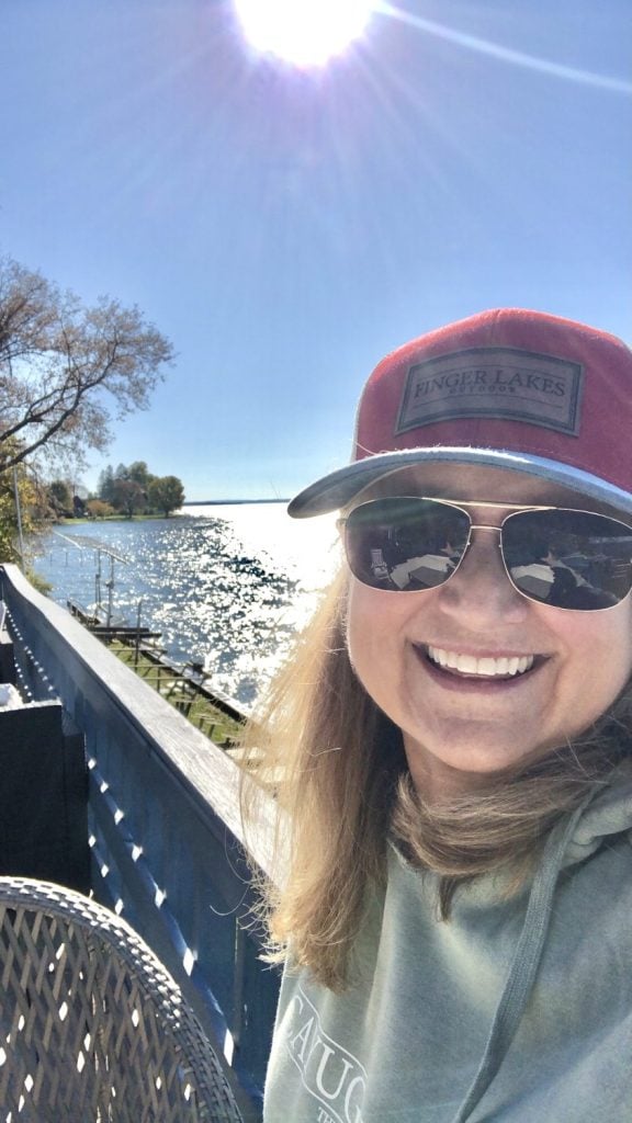Amie Freling is standing on her deck on Cayuga Lake with a finger lakes hat and smiling with the water view behind her