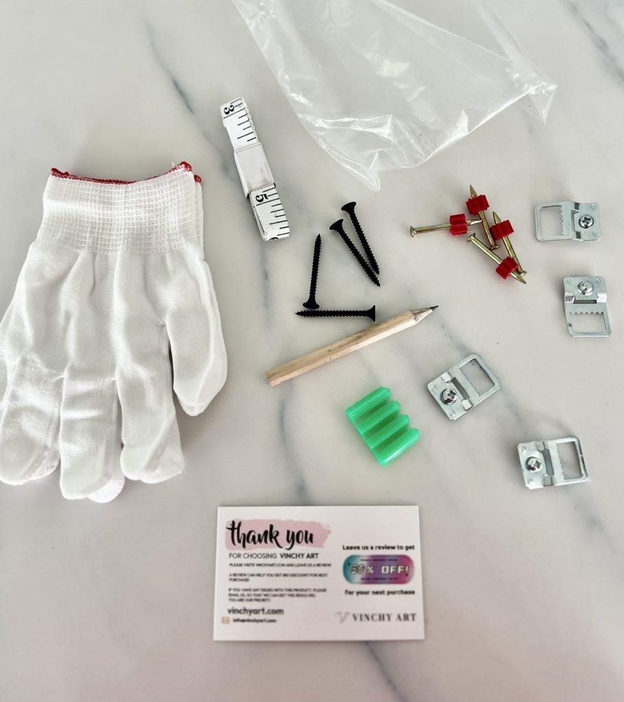 Art hanging kit from Vinchy Art laid out on a marble surface, including white gloves, screws, anchors, brackets, a pencil, measuring tape, and a thank-you card.