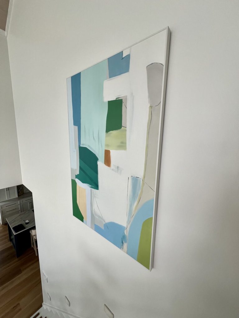 A collage of modern abstract and figurative artwork from Vinchy Art displayed in styled interiors, featuring a large blue and green abstract painting in a stairwell, additional colorful abstract pieces, and designer Amie Freling posing beside her chosen artwork.