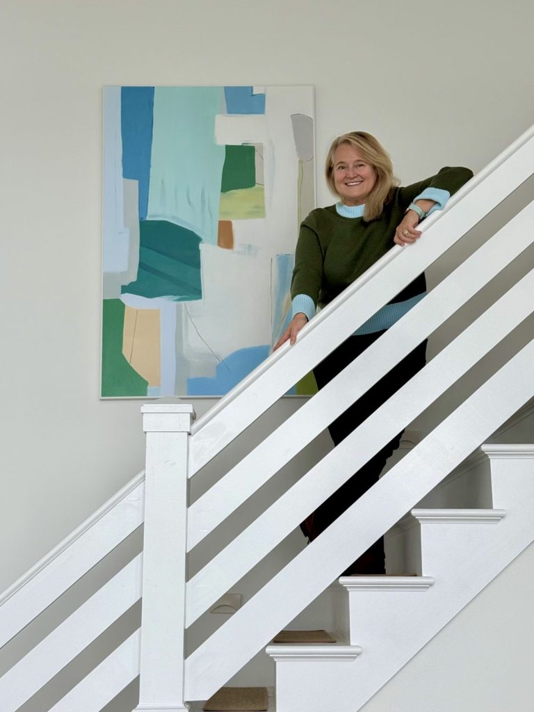 Amie Freling standing on a white staircase in her lake house, posing beside a large vertical abstract painting in shades of blue, green, and turquoise from Vinchy Art.