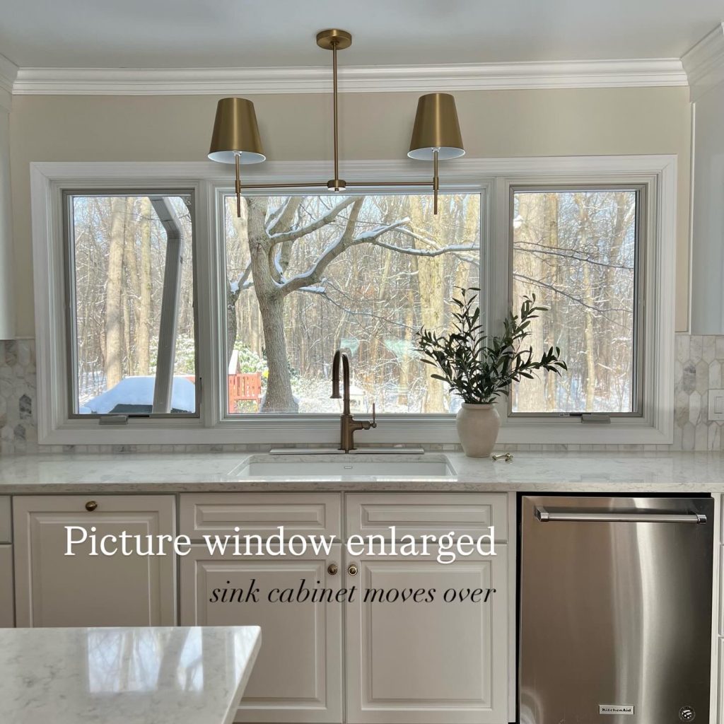 Kitchen sink area with expanded picture window, white cabinets, and brass lighting in a bright remodeled space.