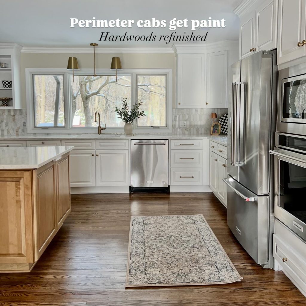 Bright renovated kitchen with white painted perimeter cabinets, stainless steel appliances, brass hardware, and refinished hardwood floors, viewed from the island toward a large window and farmhouse sink.