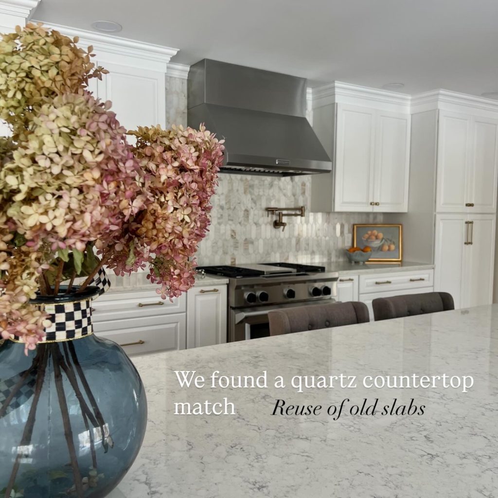 Luxury kitchen remodel with reused quartz countertops, large waterfall island, white shaker cabinets, stainless steel range and hood, champagne bronze hardware, and decorative backsplash in a bright open-concept kitchen renovation.